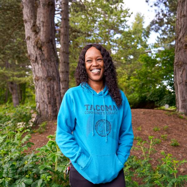A headshot of Laquida. She smiles against a wooded background, wearing a bright blue "Tacoma Community College" sweatshirt.