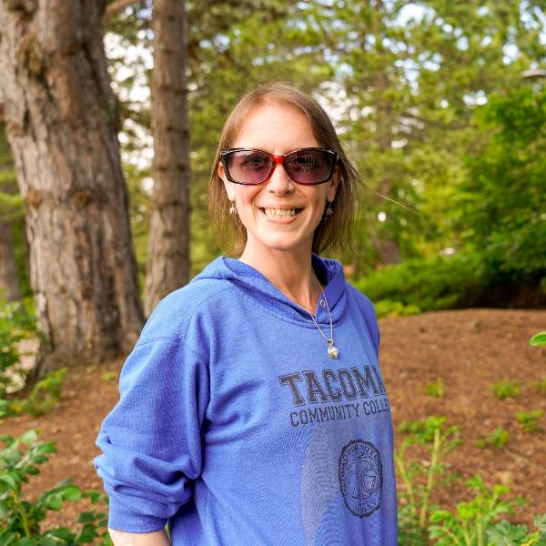 A headshot of Leigh. She smiles against a wooded background, wearing rectangular sunglasses and a royal blue "Tacoma Community College" sweatshirt.