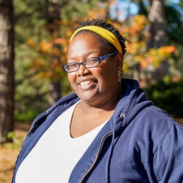 A headshot of Santina. Santina stands in front of a tan wall, smiling and wearing a blue and white striped sweatshirt.