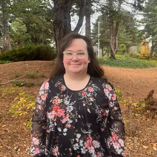 A headshot of Stevie smiling against a wooded background, and wearing a floral top