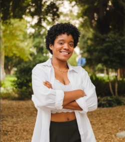 Dre stands in front of a wooded background wearing a white button-up shirt