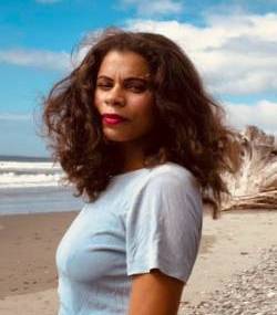 A photo of Kaela standing on the beach with wind blowing in their hair