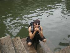 Amon Greene smiling, crouched down on steps leading to water with sunglasses and a backpack on. 