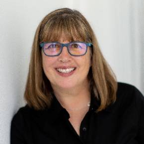 Mary Boone with shoulder-length brown hair, wearing glasses and a black shirt, standing against a white wall.