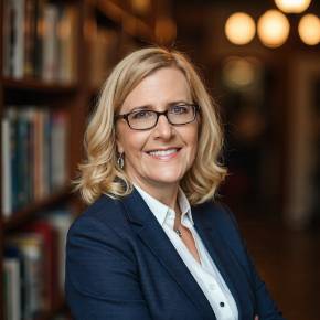 Leslie Hall with shoulder-length blonde hair in a navy blazer and white shirt, standing with arms crossed in a warmly lit library.