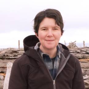 HR Heghauer in a dark jacket and plaid shirt standing outdoors with driftwood and a cloudy sky in the background.
