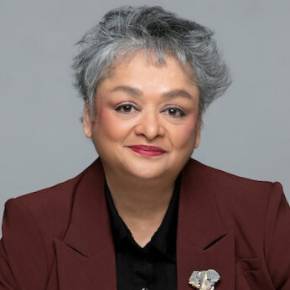 Photo of Shama Shams with short gray hair in a dark red blazer and black shirt, standing against a plain gray background.