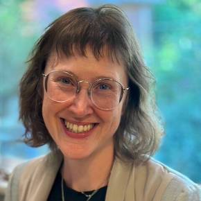 Photo of Star Wuerdemann with shoulder-length hair wearing a light jacket, dark top, and necklace, standing indoors with greenery visible through a window.