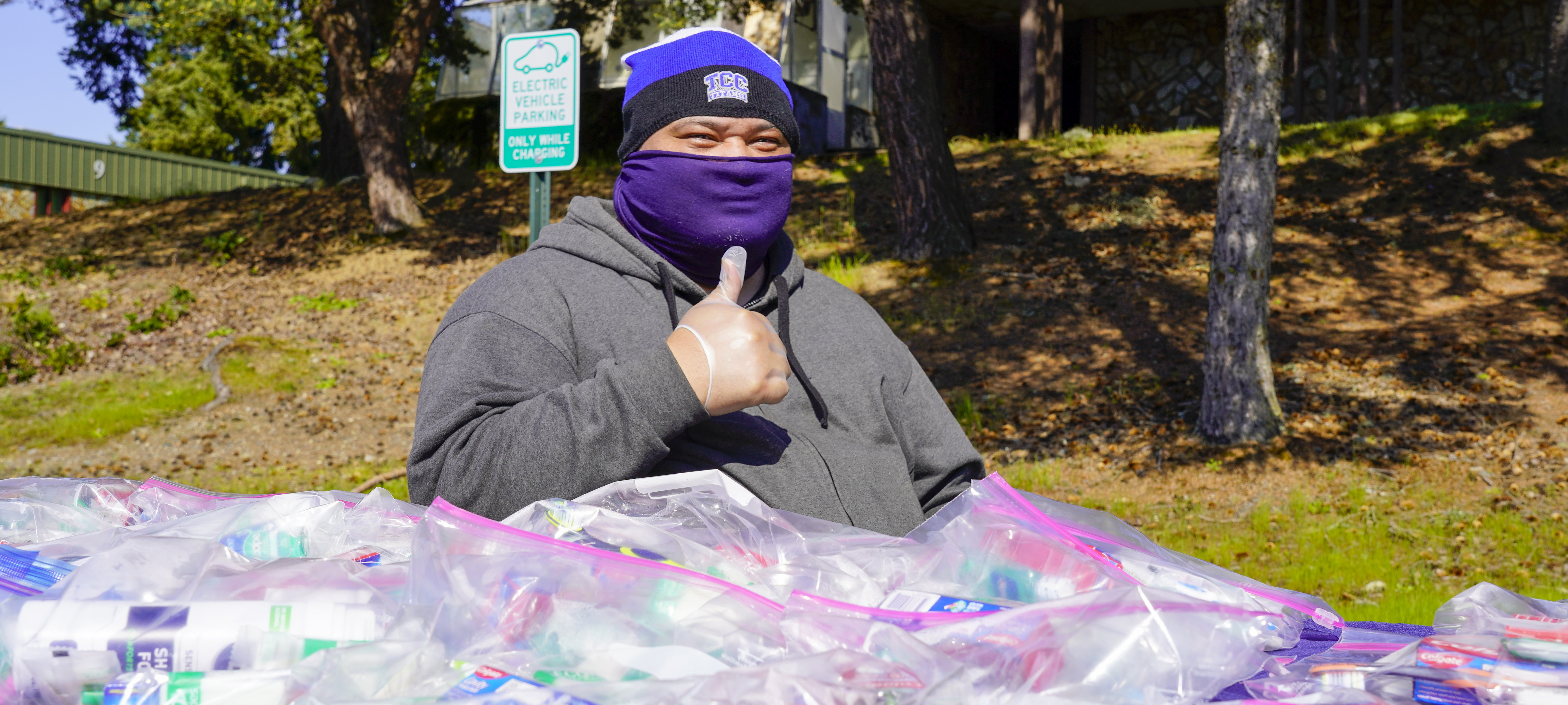 man in a mask giving a thumbs up behind a table covered with bagged supplies